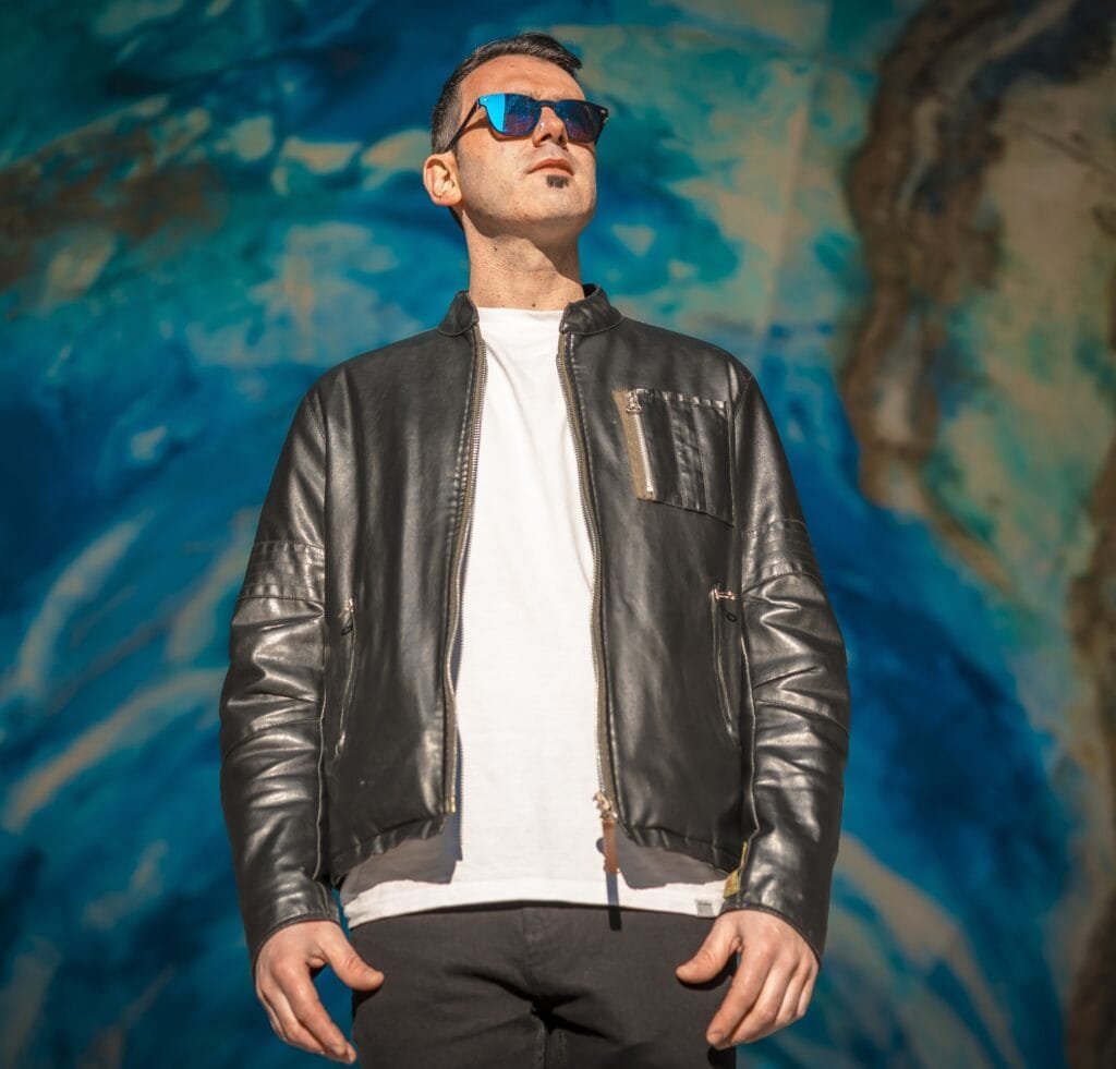 A-2 Bomber Jackets: The Perfect Blend of Rugged and Refined 4 A man wearing a black leather A-2 bomber jacket over a white T-shirt, paired with dark jeans and blue reflective sunglasses, standing outdoors in front of a colorful mural.