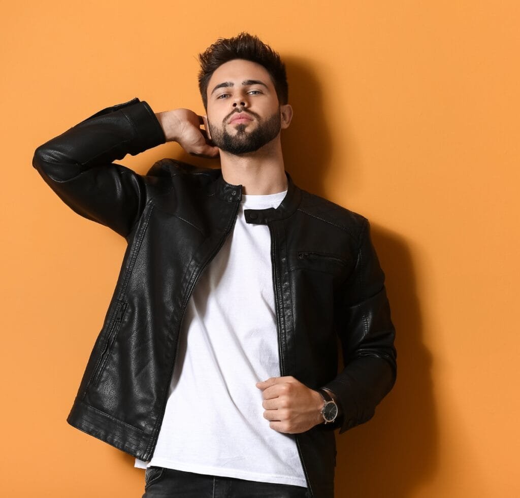 A stylish man with a trimmed beard and short dark hair posing against an orange background. He wears a black leather bomber jacket over a white T-shirt, one hand touching the back of his neck and the other resting on his jacket, with a silver wristwatch visible on his left wrist. Types of Bomber Jackets