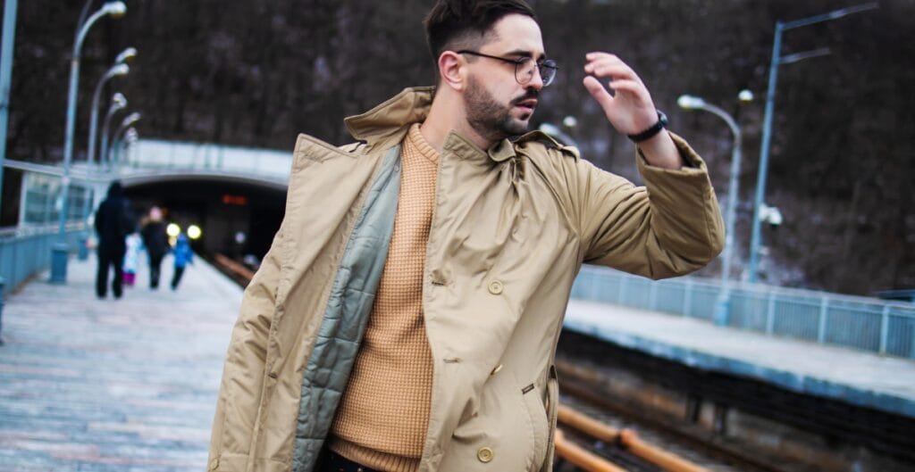 A man wearing a beige trench coat and tan sweater stands on a train platform, adjusting his eyeglasses as the wind blows his coat.