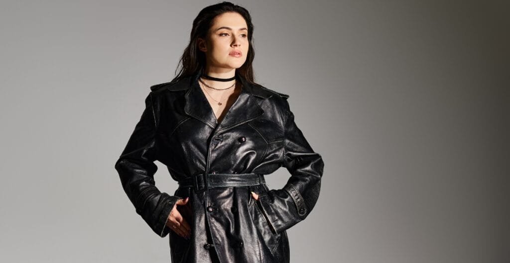 Woman wearing a black leather trench coat, standing with hands in pockets.