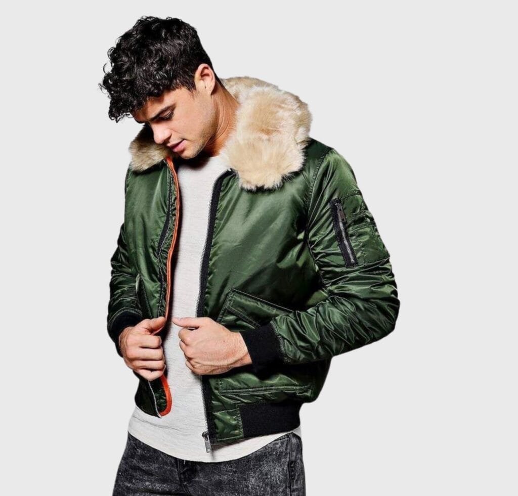 A man wearing a green bomber jacket with a beige faux fur collar, zipper front, and ribbed cuffs. He is looking down while zipping up the jacket, with a white shirt and dark jeans.