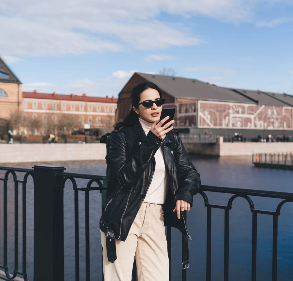A stylish woman stands by a railing overlooking a river, wearing a black leather bomber jacket, cream turtleneck, beige high-waisted trousers, dark sunglasses, and a backpack, while holding a smartphone.