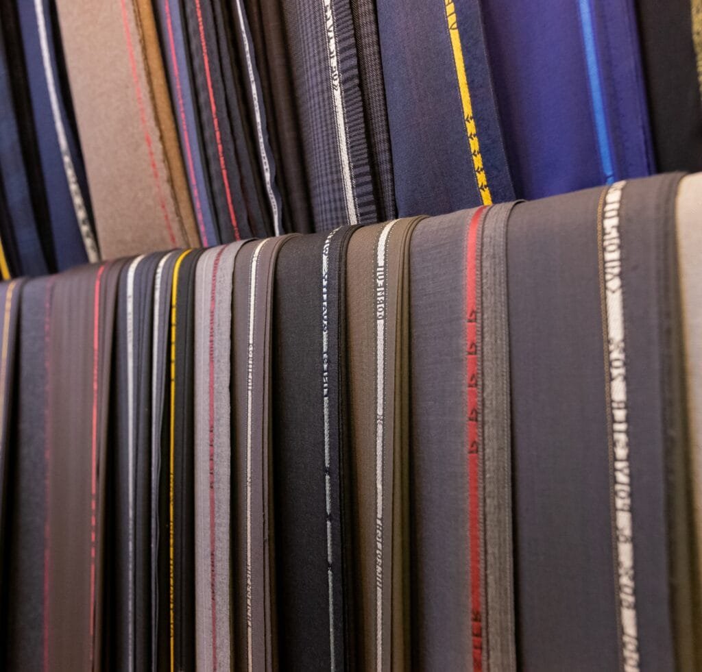 A row of folded suiting linen fabrics in various solid colors and subtle weaves, standing upright on shelves.