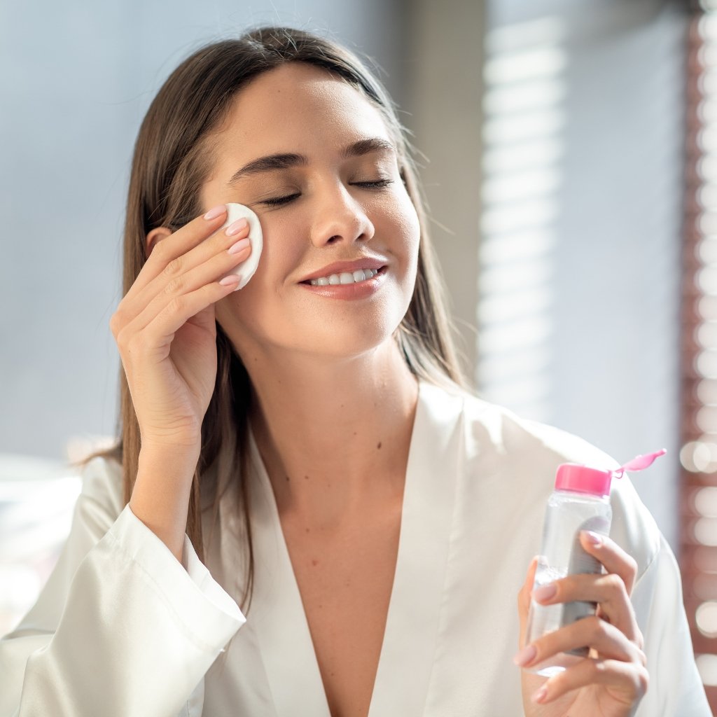 Why Supima Cotton Feels Softer, Stronger, and Smarter 22 A young woman wearing a white robe applies facial skincare with a cotton pad on her cheek, holding a clear bottle with a pink cap in the other hand, in a bright, softly lit room.