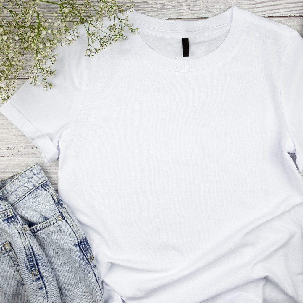 Why Supima Cotton Feels Softer, Stronger, and Smarter 16 A plain white T-shirt laid flat on a light wooden surface, with a pair of light-washed denim jeans partially visible in the bottom left corner and sprigs of small white flowers in the top left corner.