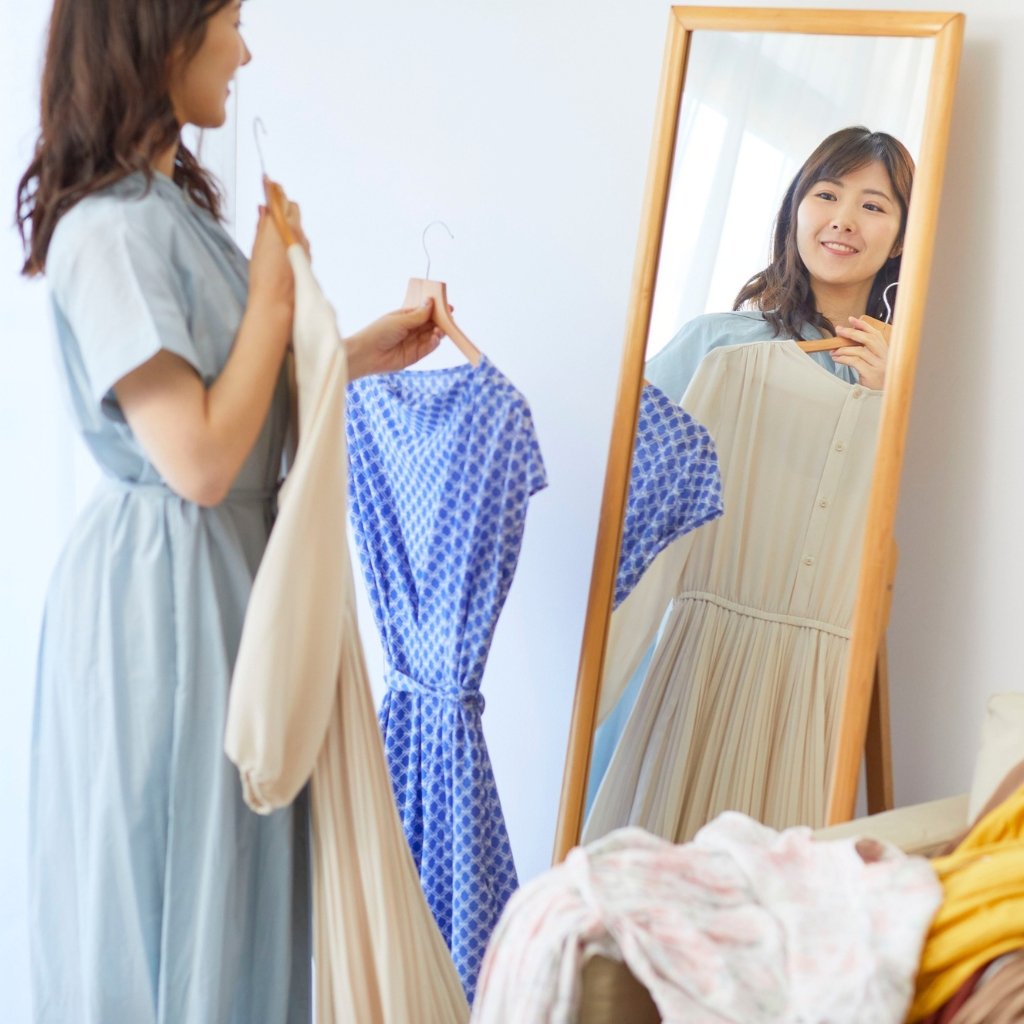Why Supima Cotton Feels Softer, Stronger, and Smarter 24 A woman stands in a bright room trying on dresses. She holds a hanger with a cream-colored dress while looking at herself in a full-length wooden-framed mirror; a blue patterned dress hangs nearby. Soft, pastel tones create a calm boutique vibe.