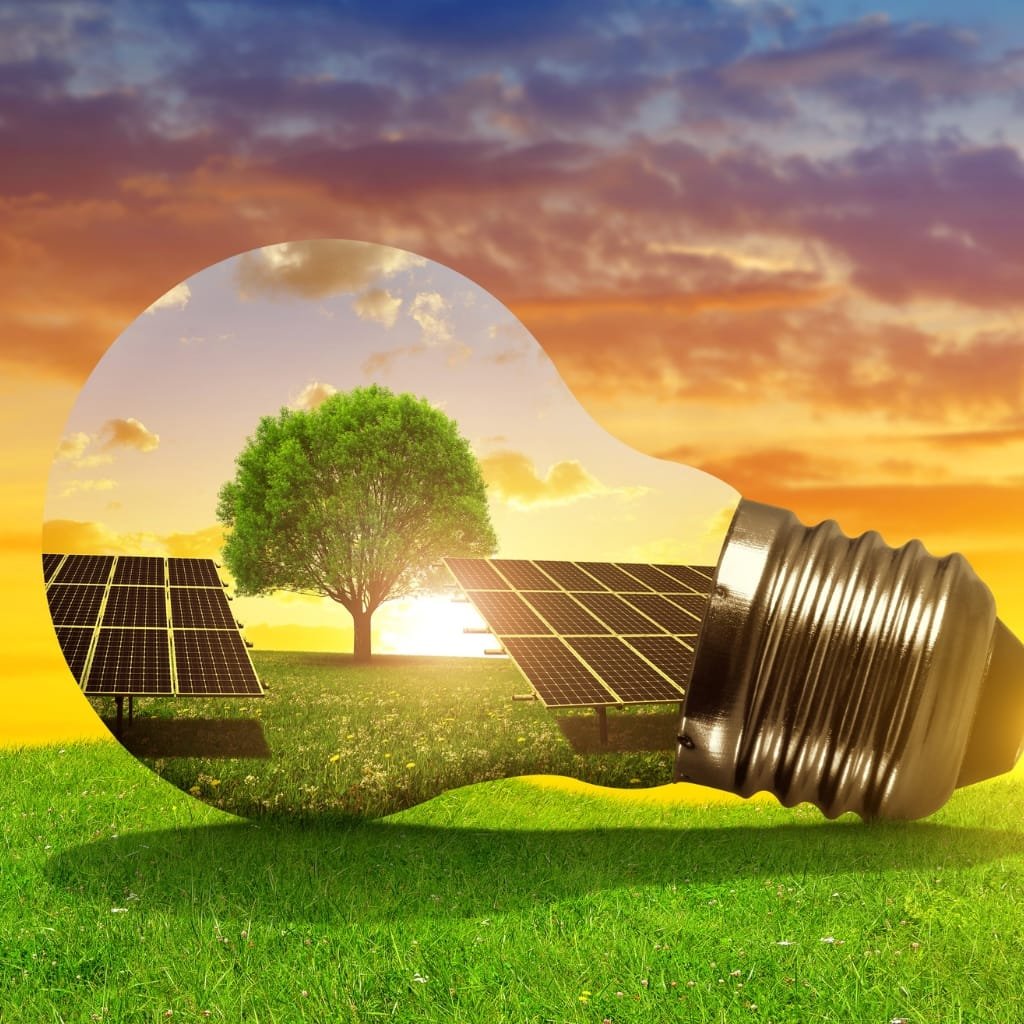 A bright light bulb lying on green grass, with a landscape inside the bulb showing solar panels, a tree, and a sunset sky.