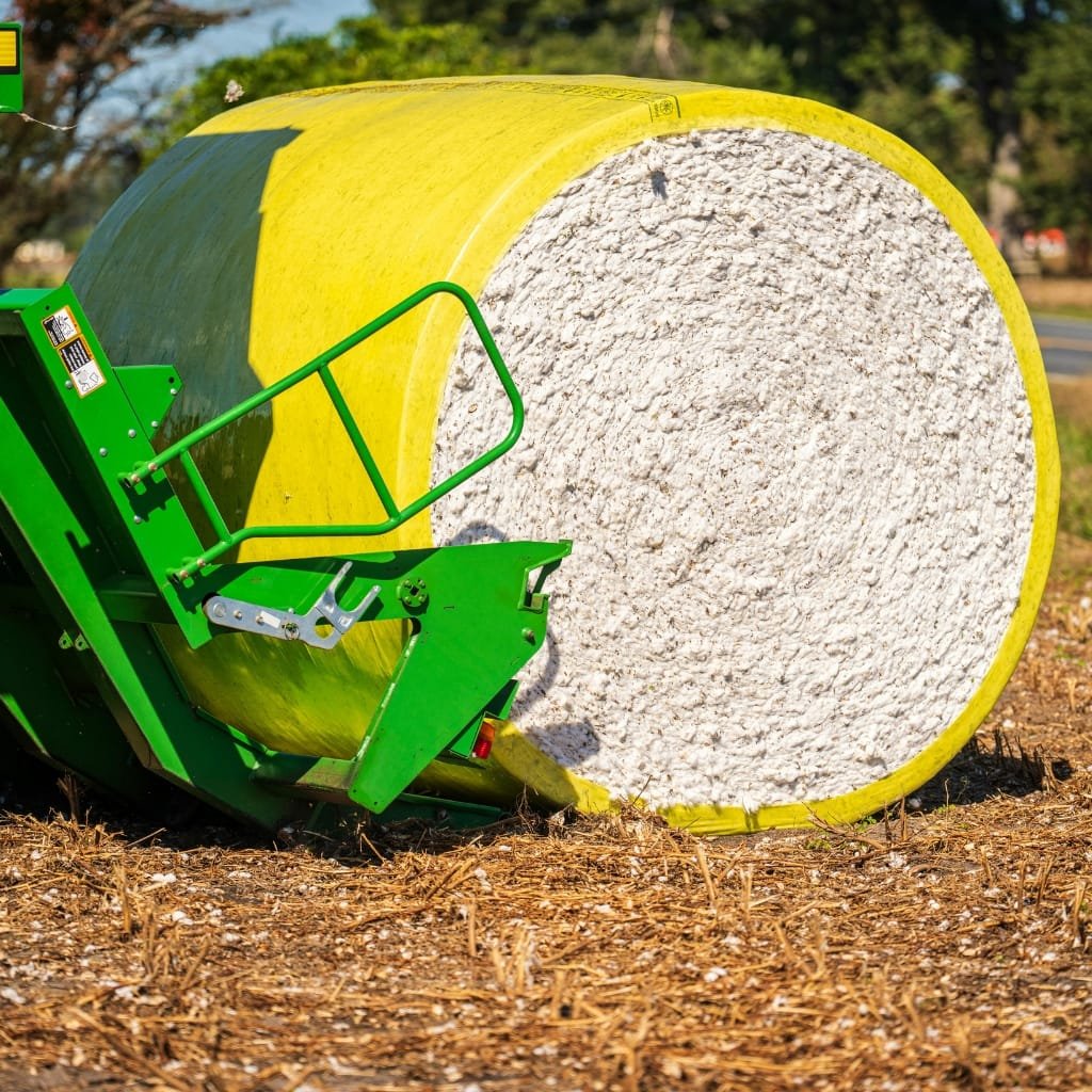 A large round bale of cotton sits next to a green farming implement in a field, illustrating modern agricultural machinery used in cotton farming.