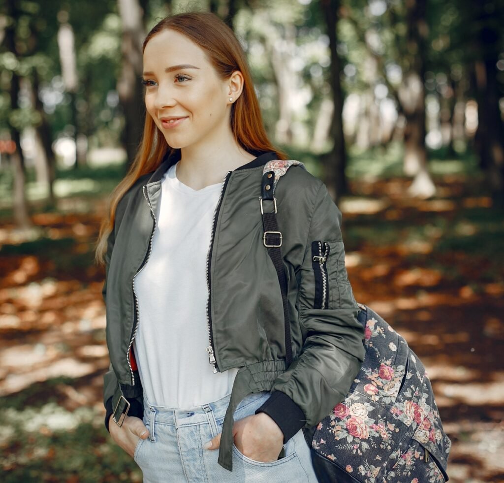 A young woman with light skin and long red hair stands outdoors in a park. She wears a military-green bomber jacket over a white T-shirt and light-wash jeans, with a floral backpack slung over one shoulder. She looks to the side with a soft smile as autumn leaves scatter on the ground.