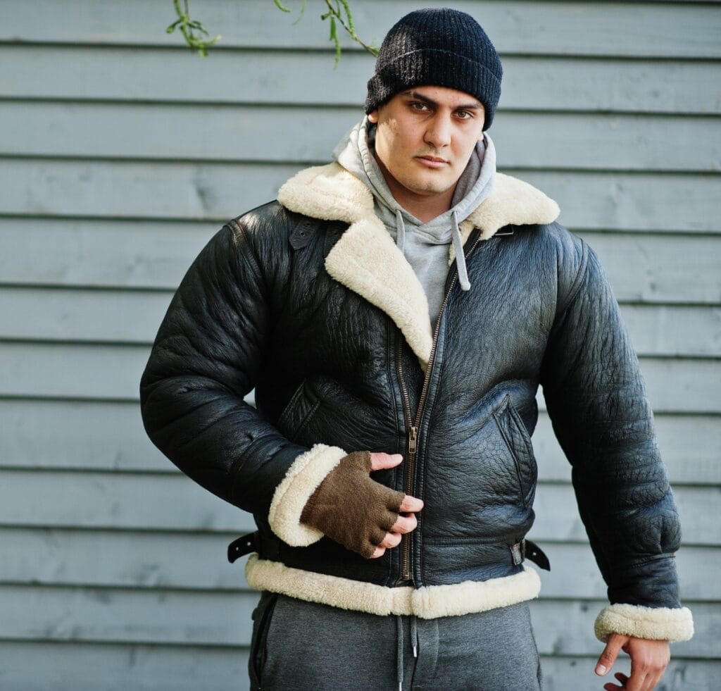 A man wearing a black leather B-3 bomber jacket with cream shearling lining and cuffs, gray hoodie, dark beanie, and brown fingerless gloves stands outdoors against a light gray wooden siding background. He looks toward the camera with a neutral expression.