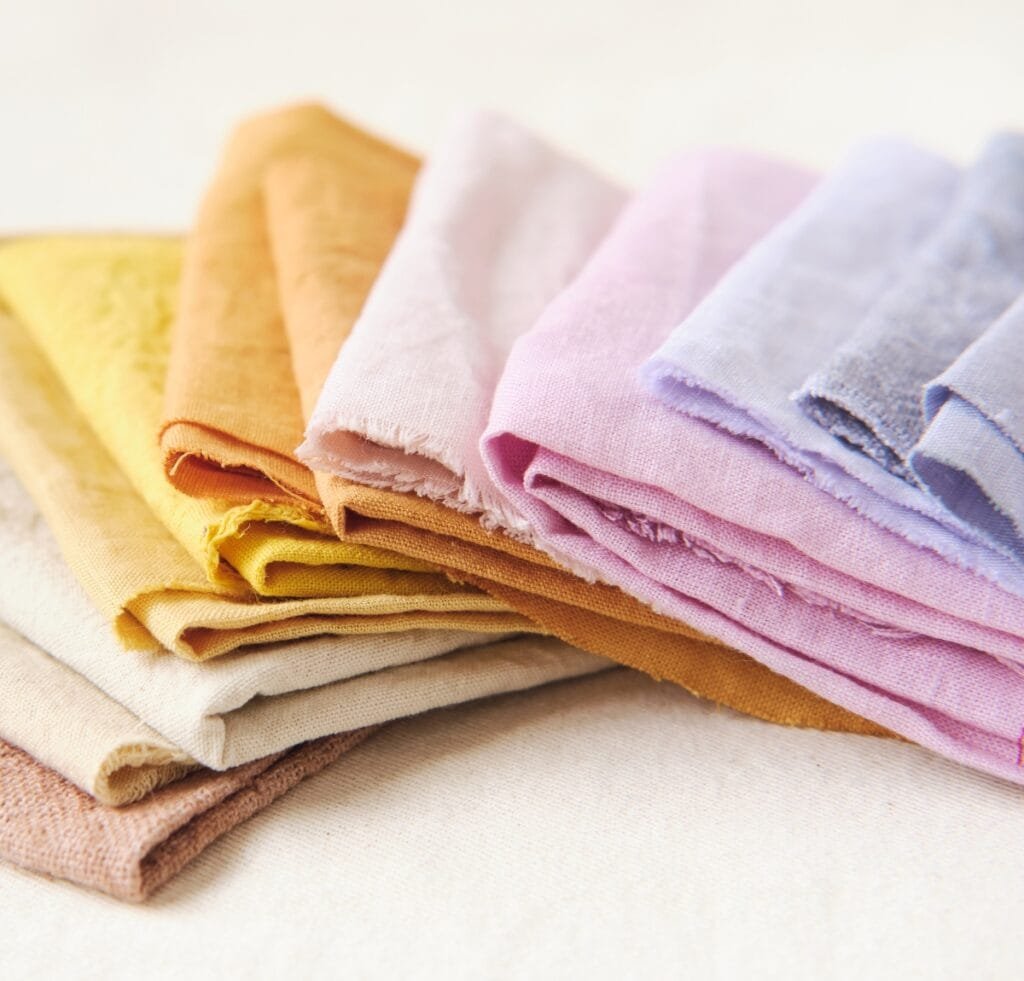 "Stack of folded linen fabrics in soft pastel colors including yellow, orange, pink, and lavender on a light surface."