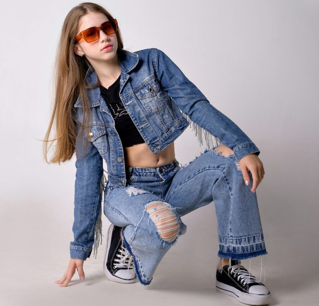 A young woman wearing a matching light-wade denim bomber jacket and jeans, styled with a black crop top, orange sunglasses, and black sneakers. She poses in a kneeling, dynamic stance against a plain background, with long straight hair.