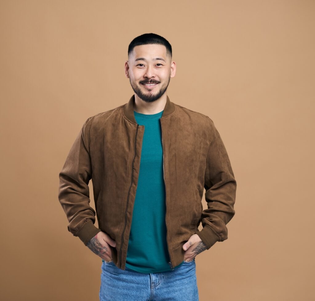 A smiling man stands against a warm beige background wearing a brown suede bomber jacket over a teal-green shirt and light blue jeans. He has short black hair and a neatly trimmed beard, with his hands casually tucked into the jacket pockets.