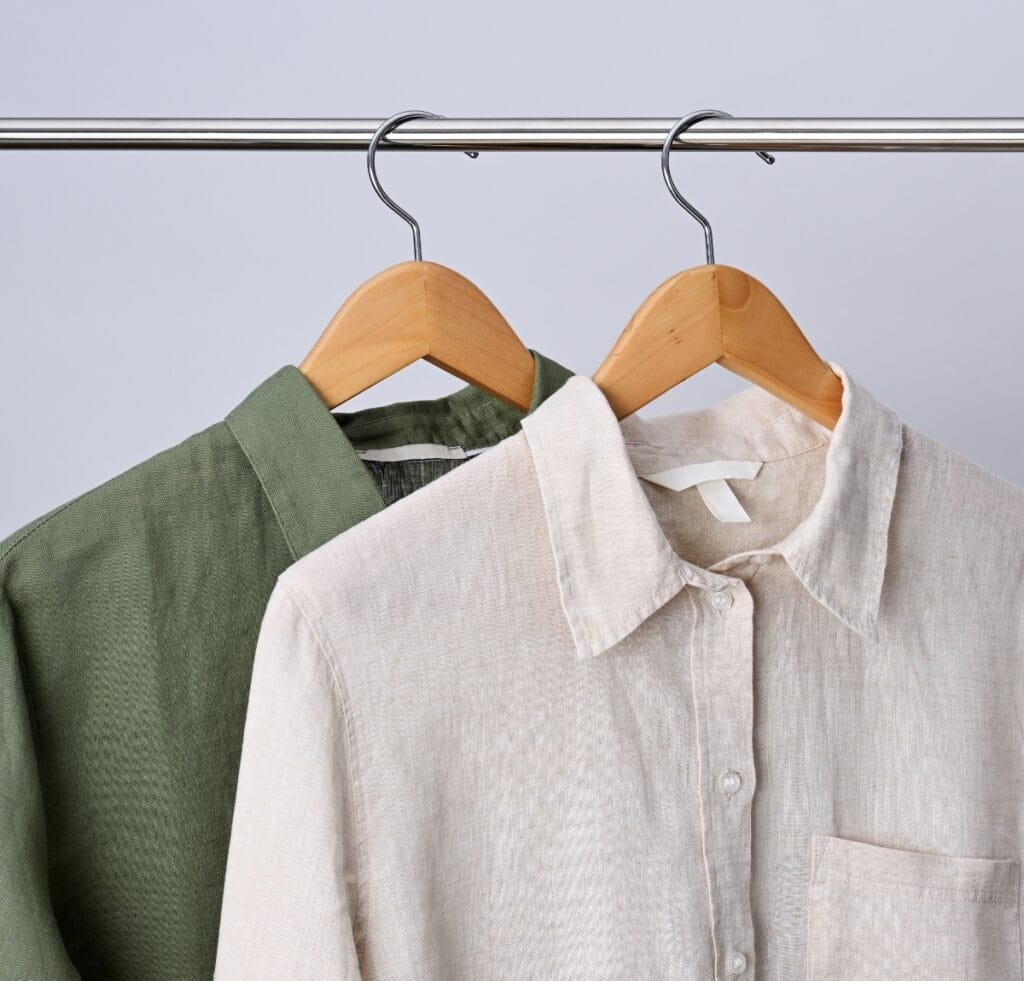 Linen Outfit Fabric Essentials: Timeless, Lightweight, Versatile 7 “Two linen shirts on wooden hangers hanging from a metal clothing rod. The front shirt is light beige with a collar and pocket; the shirt behind is olive green.” Linen Outfit Fabric
