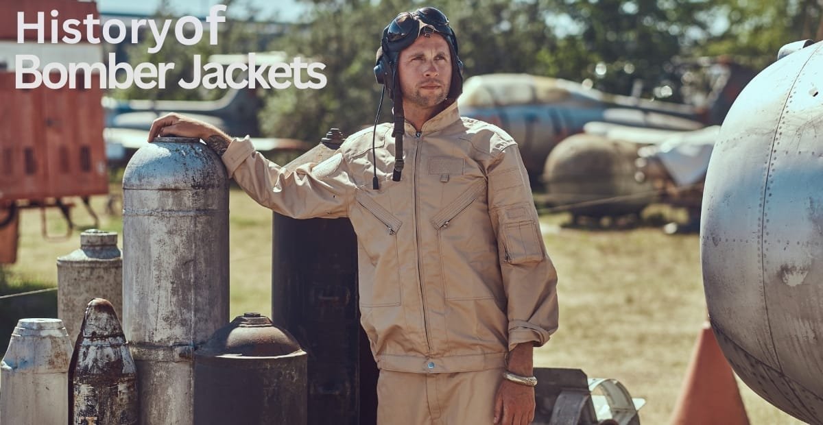 "A man in a tan bomber jacket wearing a flight helmet rests his hand on a metal canister in an outdoor setting with vintage aircraft parts in the background."