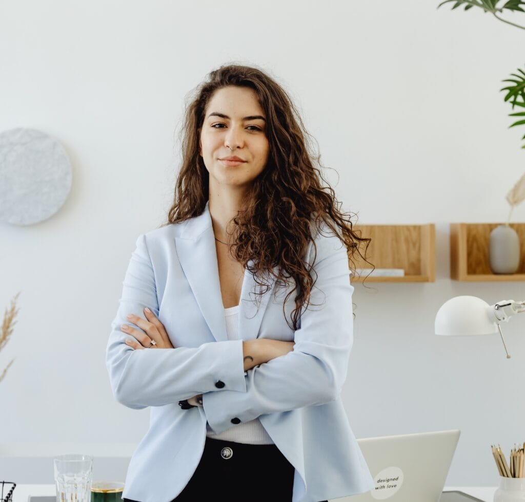 Linen Outfit Fabric Essentials: Timeless, Lightweight, Versatile 11 A young woman with curly hair stands confidently in a light blue blazer, arms crossed, in a bright, modern office.