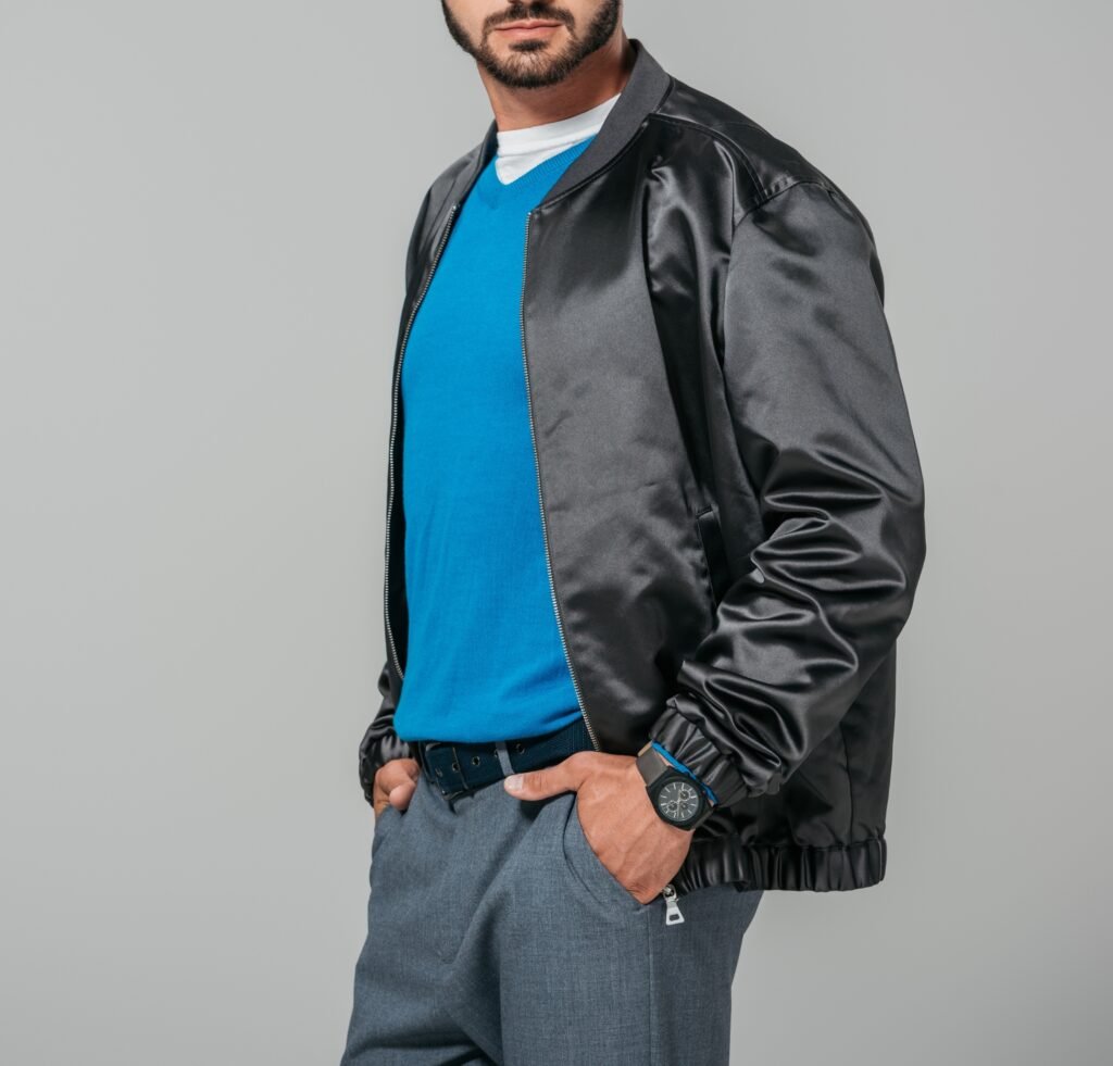 A man wearing a sleek black satin bomber jacket over a bright blue polo or t-shirt, paired with gray tailored trousers. He has a black belt and a dark wristwatch, with hands in his pockets, posing against a neutral backdrop.