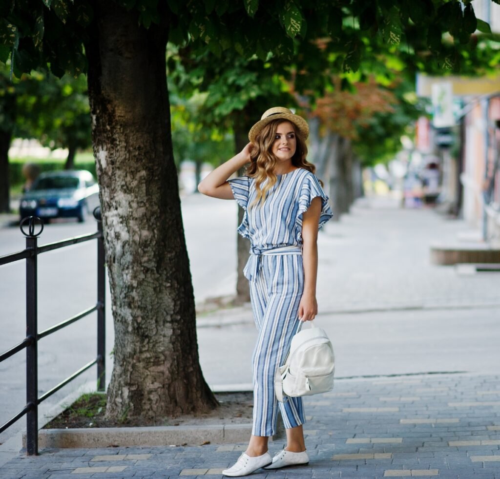 Linen Outfit Fabric Essentials: Timeless, Lightweight, Versatile 10 A stylish young woman wearing a blue-and-white striped linen jumpsuit, white sneakers, a straw hat, and carrying a white backpack, standing on a sidewalk beside a tree in an urban street setting.