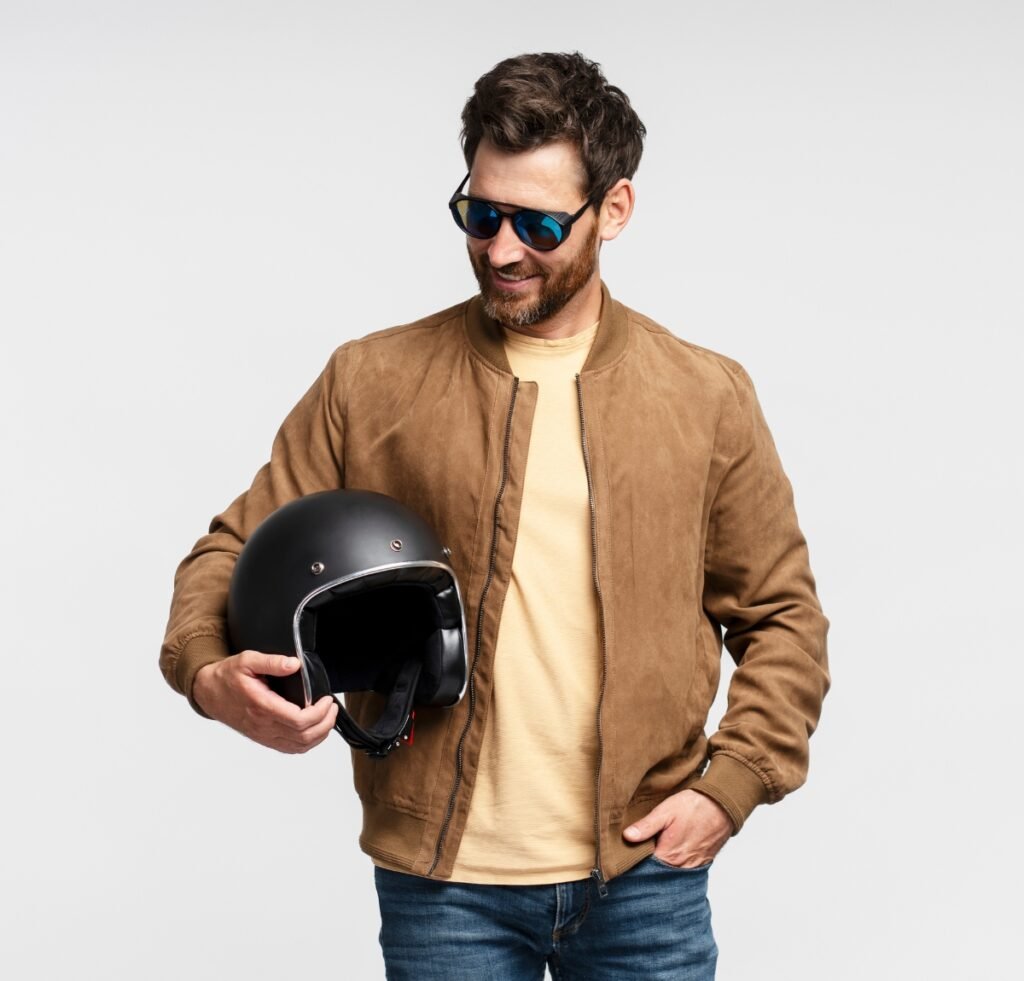 A stylish man wearing a light brown suede bomber jacket over a beige T-shirt and blue jeans, holding a black open-face helmet while wearing reflective sunglasses.