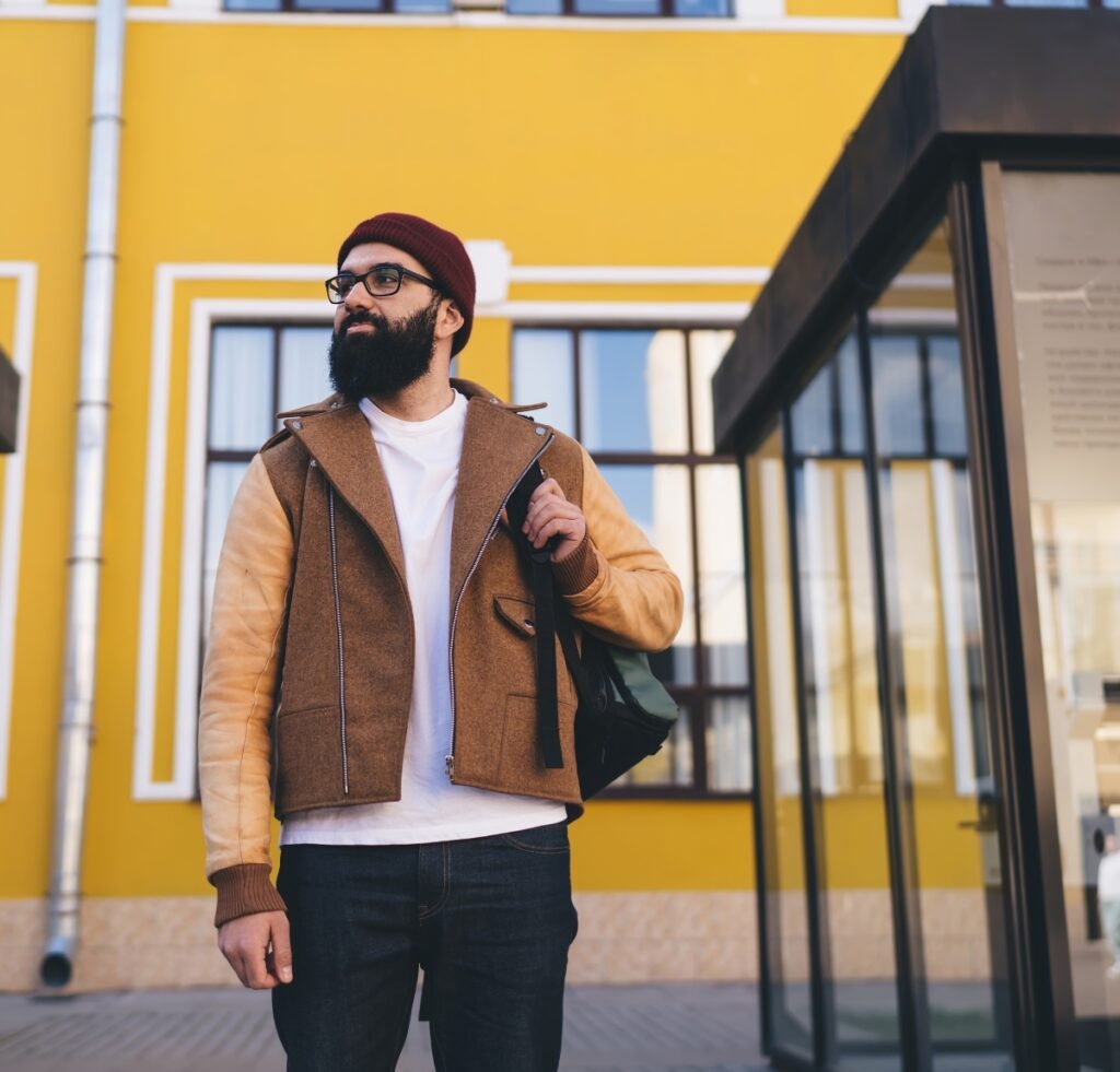 "A stylish man with a beard and glasses wearing a two-tone suede bomber jacket, white t-shirt, dark jeans, and a maroon beanie, standing outdoors in front of a bright yellow building and a modern glass entrance."