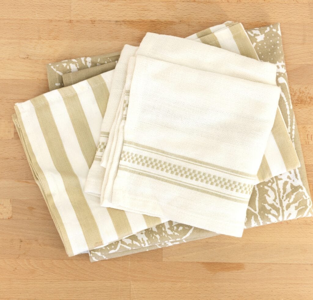 A neatly folded stack of linen towels in natural, neutral tones (beige, cream, and light tan) featuring striped and decorative woven patterns, placed on a light wooden surface.