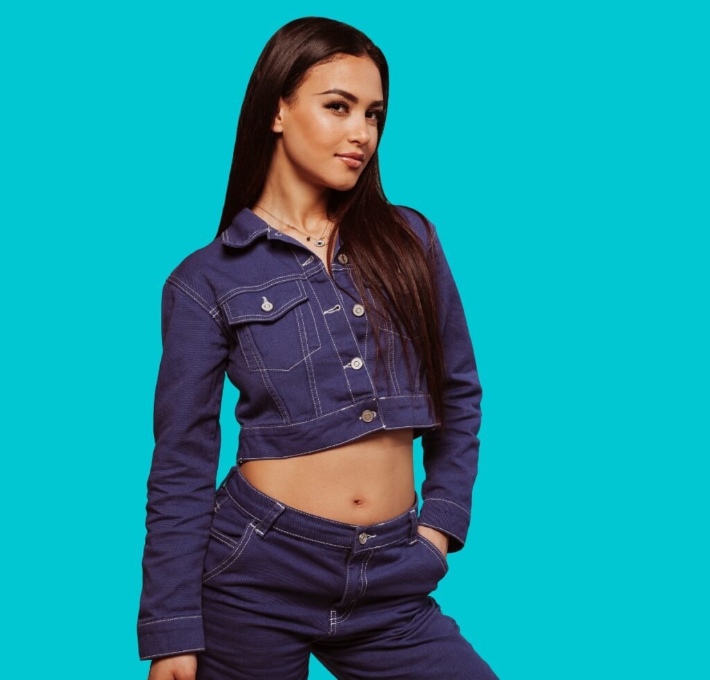 "Young woman wearing a cropped denim bomber jacket and matching high-waisted jeans, standing against a solid cyan background. She has long dark hair and a confident pose with one hand in her pocket."