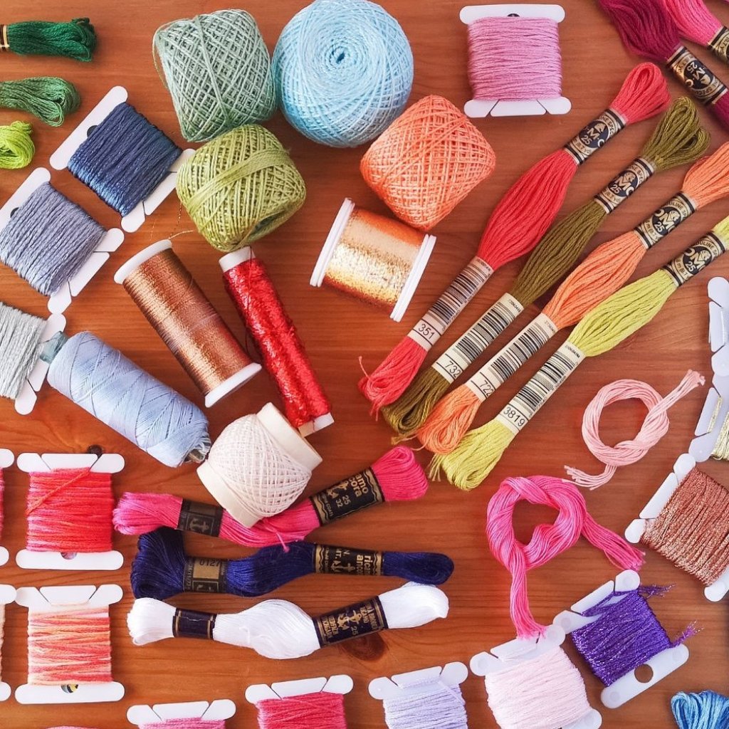 A colorful assortment of embroidery threads, floss, and yarns in various colors and textures arranged on a wooden surface.