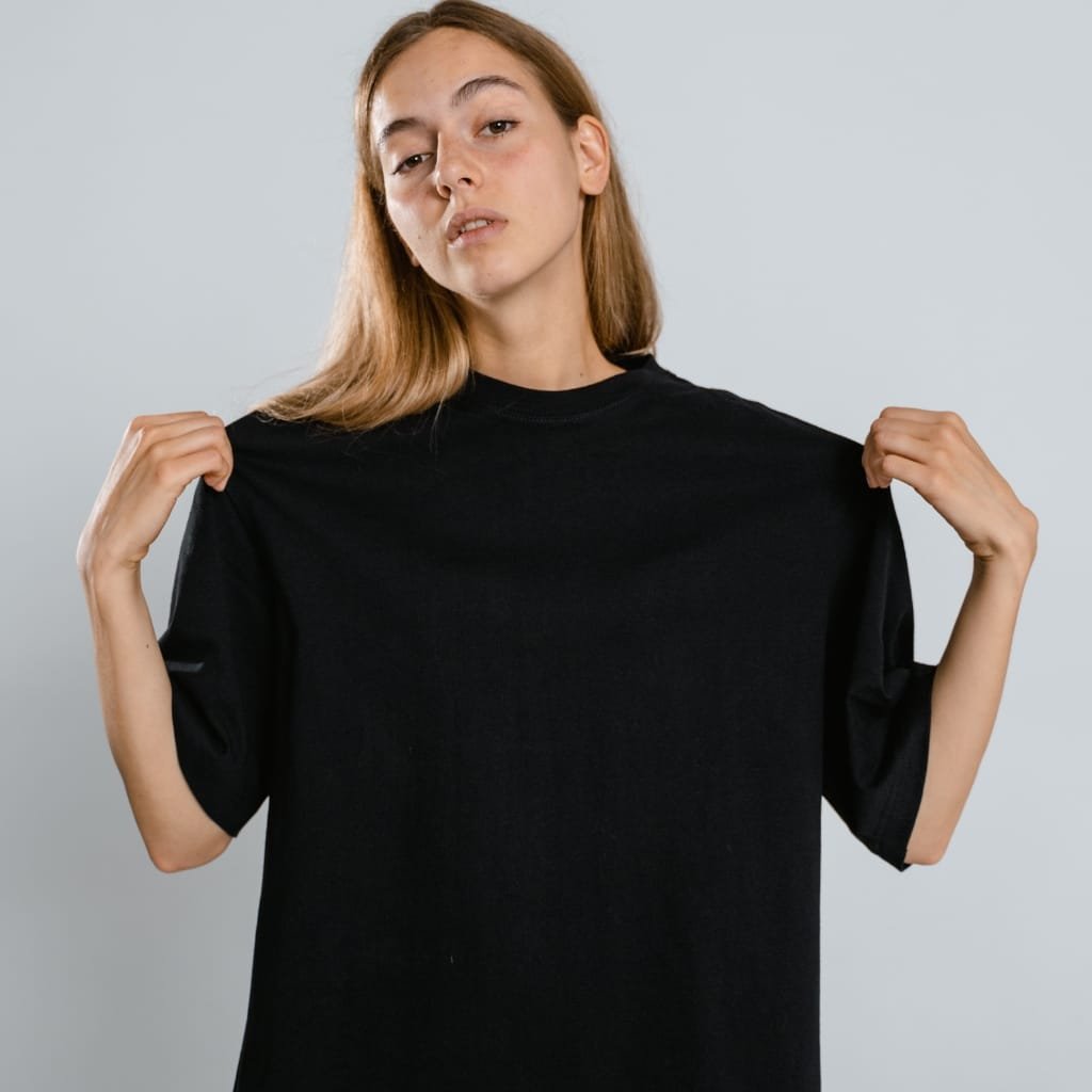  A young woman with light brown hair pulls on the sleeves of an oversized black T-shirt, facing the camera against a light gray background.