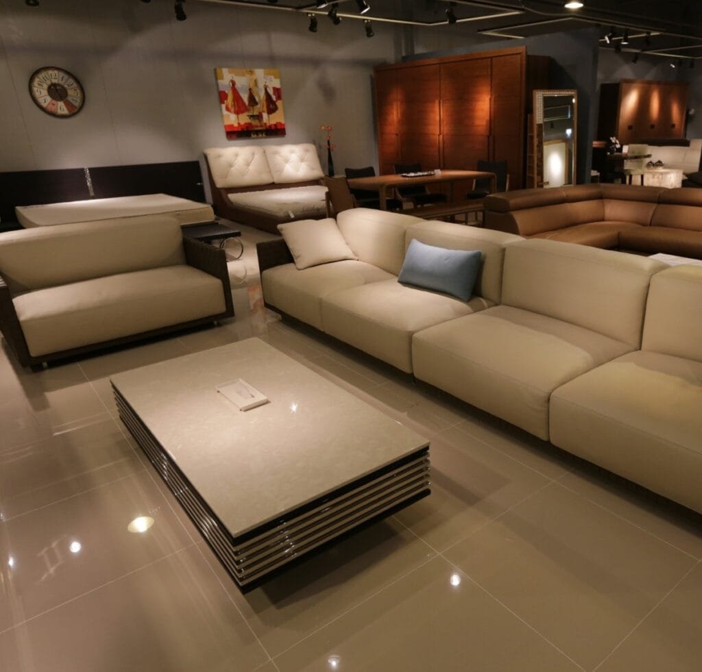 A spacious showroom of modern upholstered furniture. A large cream-colored sectional sofa with a matching loveseat and a light blue throw pillow sits around a low, rectangular coffee table with a layered, glossy stone top. In the background are multiple beds, a brown leather sofa, and dark wood cabinetry, under track lighting and a neutral gray wall.