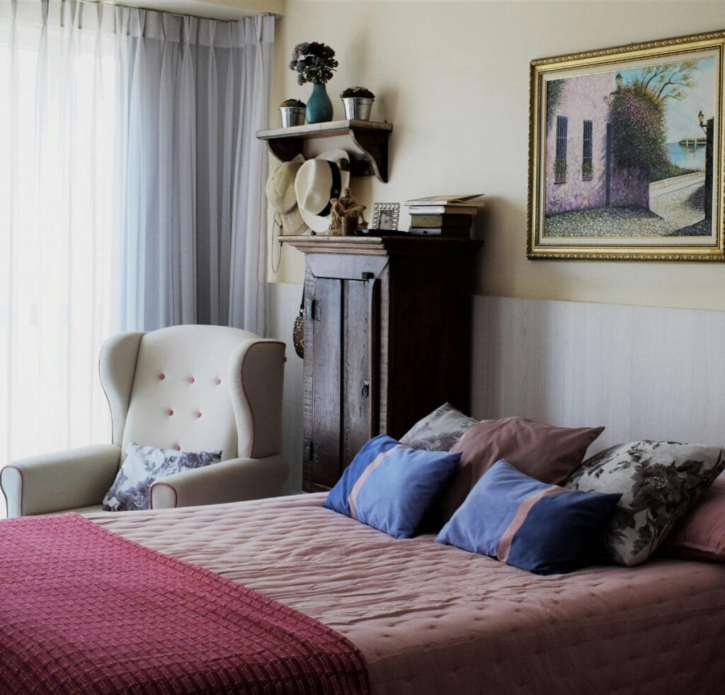 A cozy bedroom featuring a pink upholstered bed with a tufted headboard, plush blue and floral accent pillows, and a textured pink throw. Beside the bed is a cream-colored, high-back armchair with a floral cushion. A dark wooden armoire stands against the wall, topped with stacked books and hats. The wall showcases a framed landscape painting, and sheer curtains diffuse soft natural light through a large window. Hebei Linen Fabric