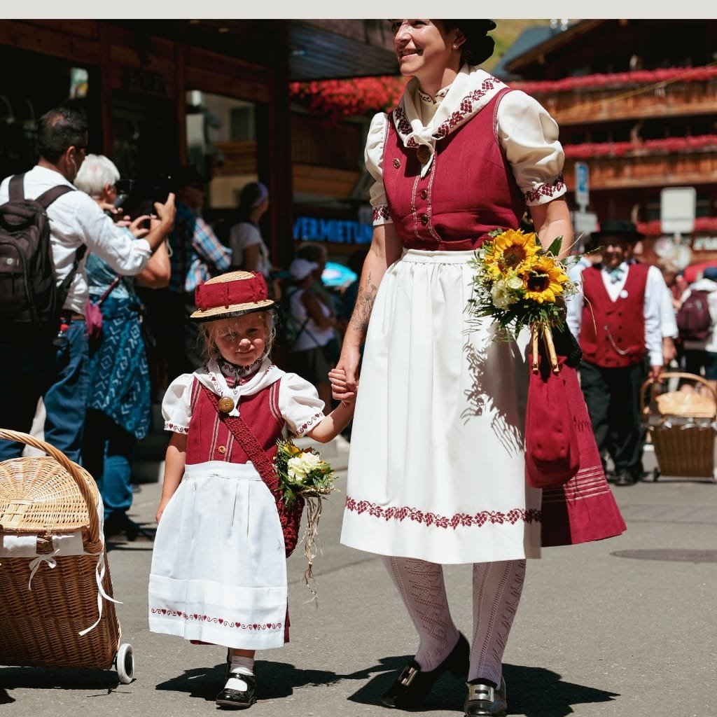 A smiling woman and a small girl dressed in traditional Bavarian-style outfits holding hands at a festival. The woman wears a burgundy dirndl with a white apron, bouquet in hand; the child wears a matching burgundy vest and hat, carrying a small bouquet. They stand among onlookers with photographers and baskets in the background.

