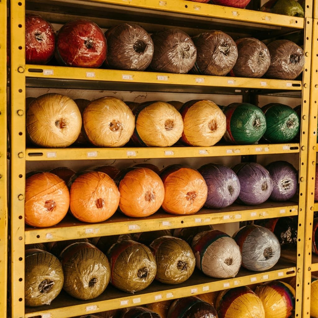 Rows of colorful yarn skeins wrapped in plastic on metal shelves in a warehouse, arranged by color from warm to cool tones.