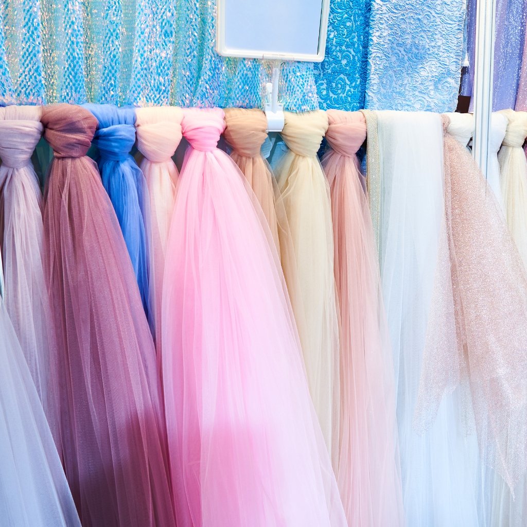 Rows of colorful crushed organza gowns or fabrics tied at the top, hanging in a pastel display with blues, pinks, purples, and creams