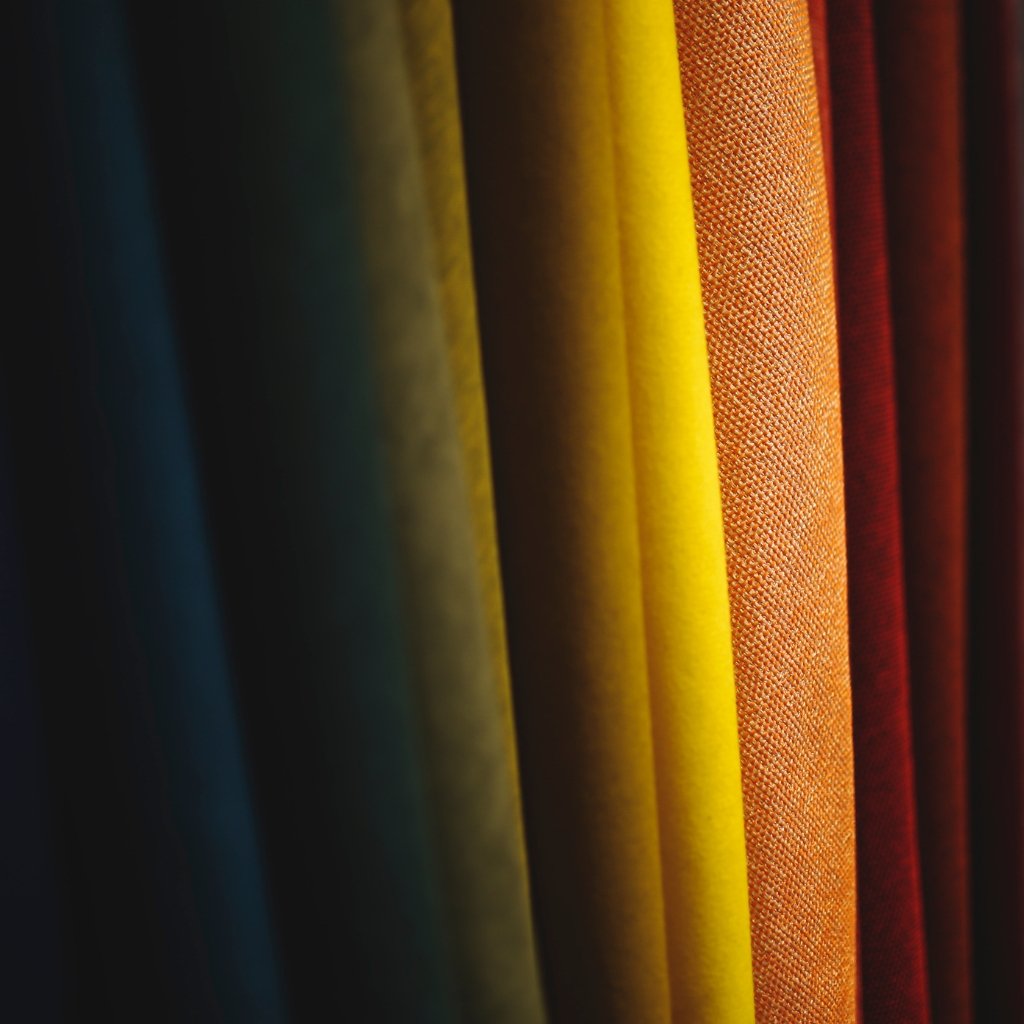 Close-up of vertical fabric rolls in a gradient of colors from dark blue to bright yellow, orange, and red. Batiste cotton fabric