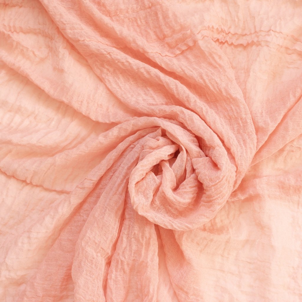 A close-up view of soft, peach-pink fabric twisted into a circular knot, showing gentle folds and a textured, airy weave.
