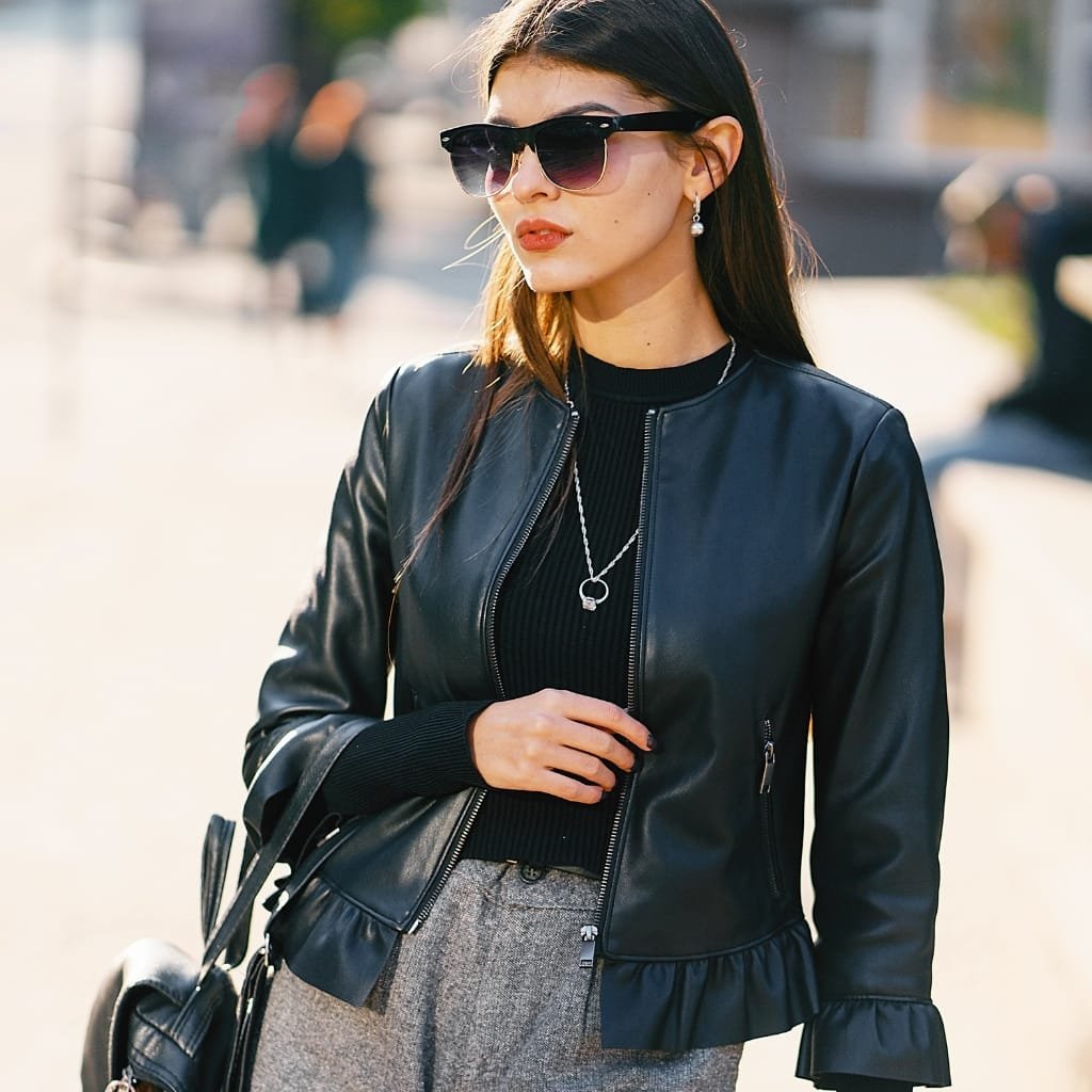 A stylish woman walking outdoors wears a fitted black leather blazer with a zippers and a ruffled hem, paired with a black knit top, gray tailored trousers, dark sunglasses, and a silver pendant necklace. She carries a black bag and accessorizes with pearl earrings, exuding a chic, modern subway-street style.