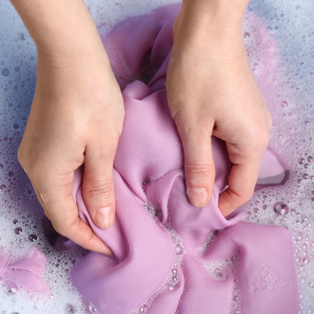 Close-up of hands wringing a piece of lavender-colored fabric in soapy water, bubbles surrounding the cloth. sateen cotton fabric