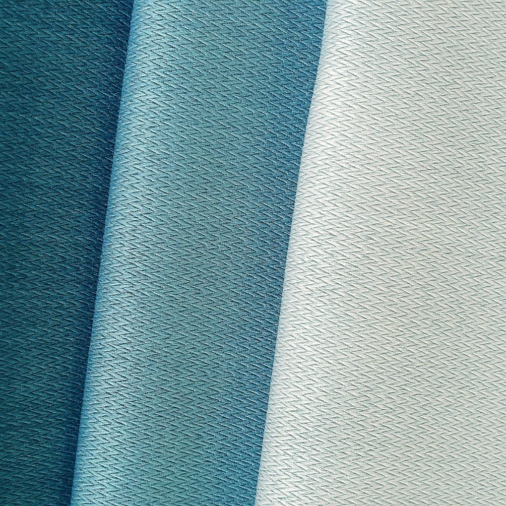 A close-up view of three rolled fabrics in varying textures and colors: a deep teal roll on the left, a slightly lighter teal in the middle, and a pale gray-beige roll on the right. All fabrics show a fine, sheer weave with a subtle crisscross pattern.
