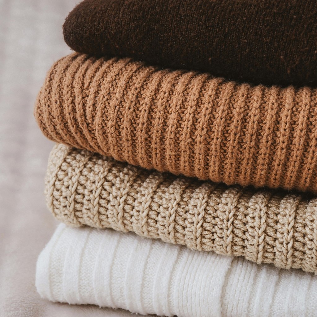 A stack of folded rib-knit cotton sweaters in shades of brown and cream, showing thick, vertical ribbed knit pattern. Rib knit cotton fabric