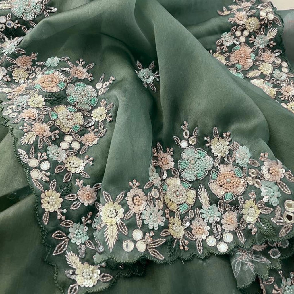 A close-up view of a soft green organza fabric adorned with dense, pastel-colored floral embroidery. The delicate flowers are created with tiny beads and sequins, forming a decorative border along the edge of the fabric.