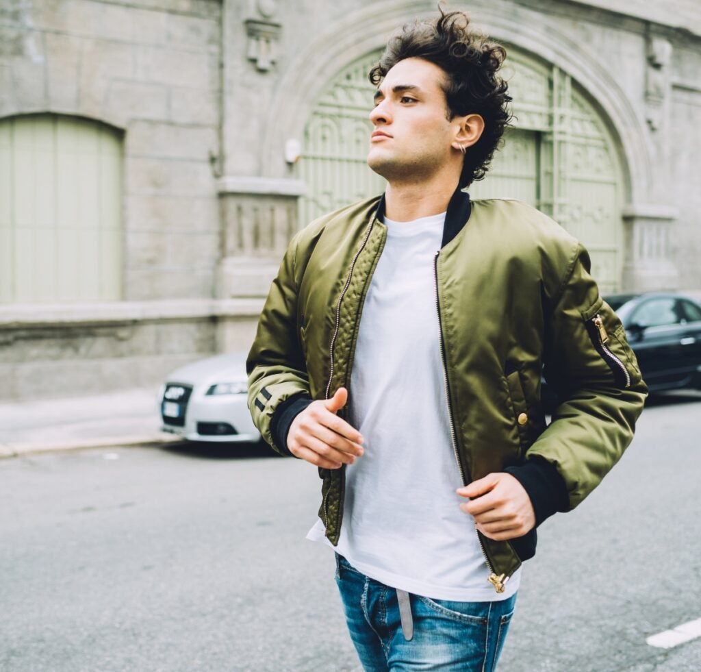 A young man wearing an olive green bomber jacket over a white t-shirt and blue jeans runs down a city street with stone buildings and a parked car in the background.