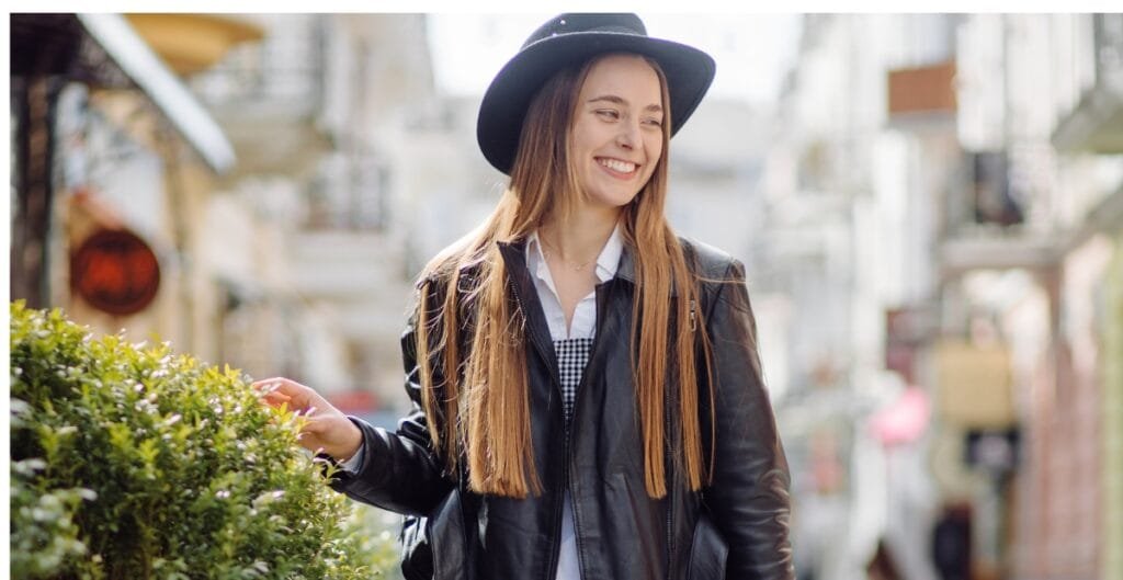 "Smiling woman wearing a black hat and leather jacket stands on a sunny street, touching a green hedge."
