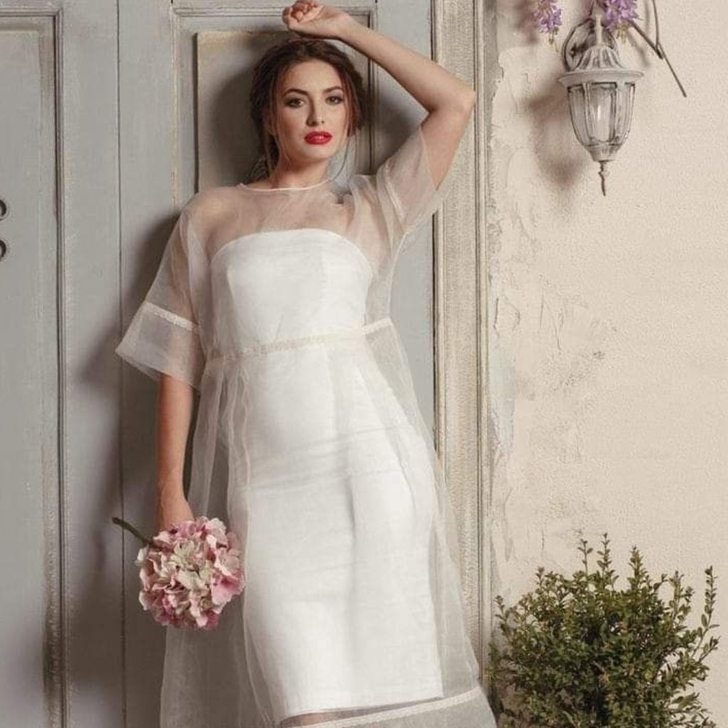 A stylish woman wearing a white strapless dress with a sheer, loose-fitting matte organza overlay. She holds a pink bouquet and stands in front of a light-colored vintage door, with a small hanging lantern and greenery visible nearby.