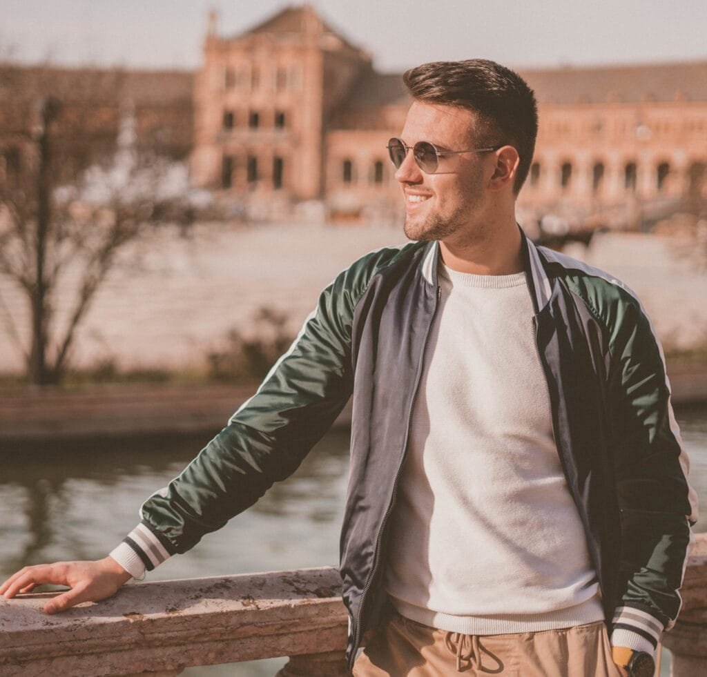 "A stylish man wearing a green and black bomber jacket over a white sweater, standing outdoors by a railing near a river. He wears sunglasses and smiles, with a historic building in the background." The History of Bomber Jackets