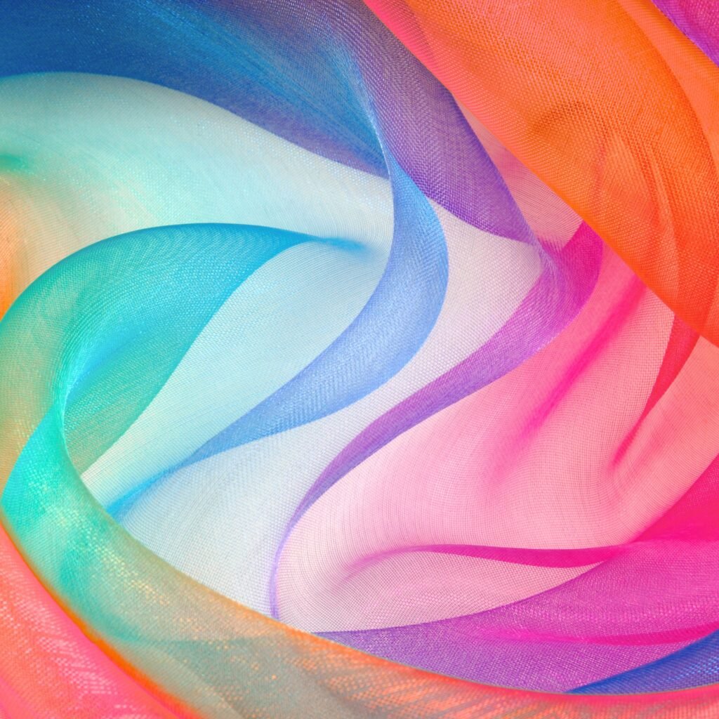 A vibrant close-up swirl of crystal organza fabric in rainbow hues, showing sheer, shimmering layers of blue, teal, pink, purple, orange, and pink arranged in soft folds and curves.