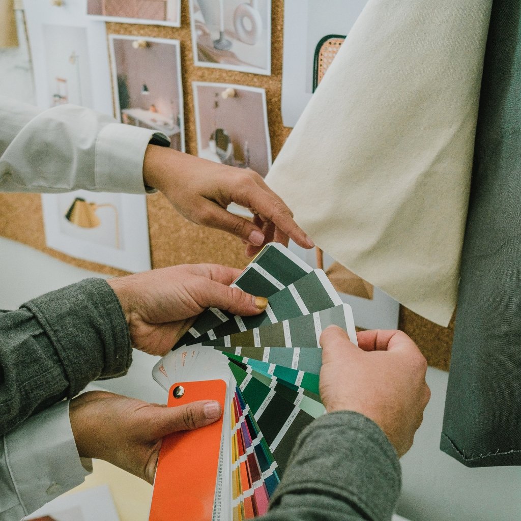 A close-up of two designers comparing color swatches. One person’s hand holds a fan of fabric color swatches in shades of green and teal, while the other points at them. A corkboard with mood boards and fabric samples is visible in the background.