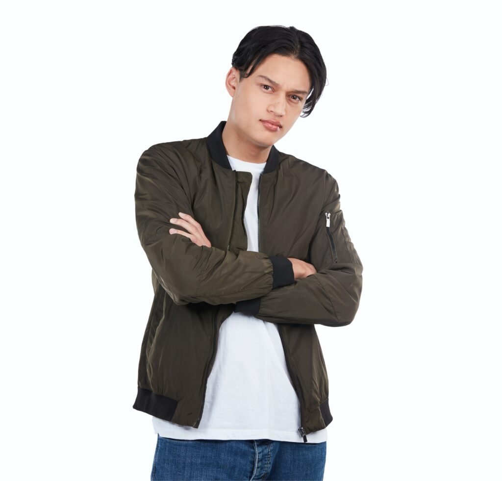 "A young man with arms crossed wearing an olive-green slim-fit bomber jacket over a white t-shirt and blue jeans, posing against a plain light background."