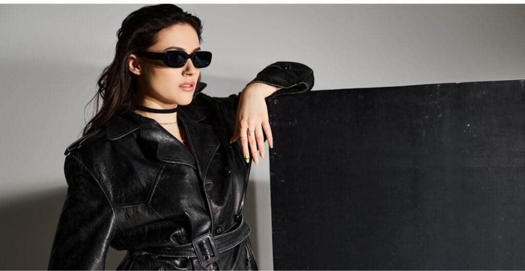 "Woman wearing a black leather trench coat and dark sunglasses, leaning on a large black panel against a light gray background."

