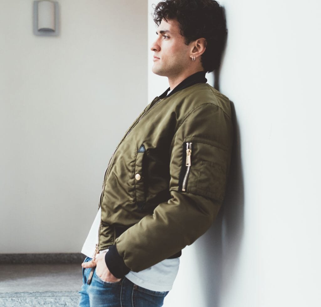 A man wearing an olive MA-1 bomber jacket leans against a white wall, hands in pockets, looking to the side.