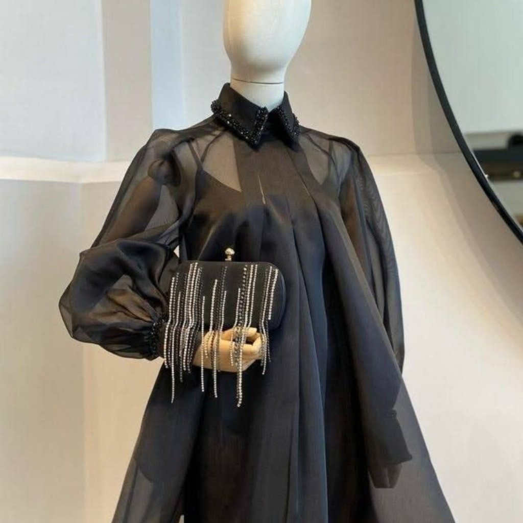 A mannequin dressed in a sheer matte black organza blouse with puff sleeves, holding a black beaded clutch adorned with dangling chains.